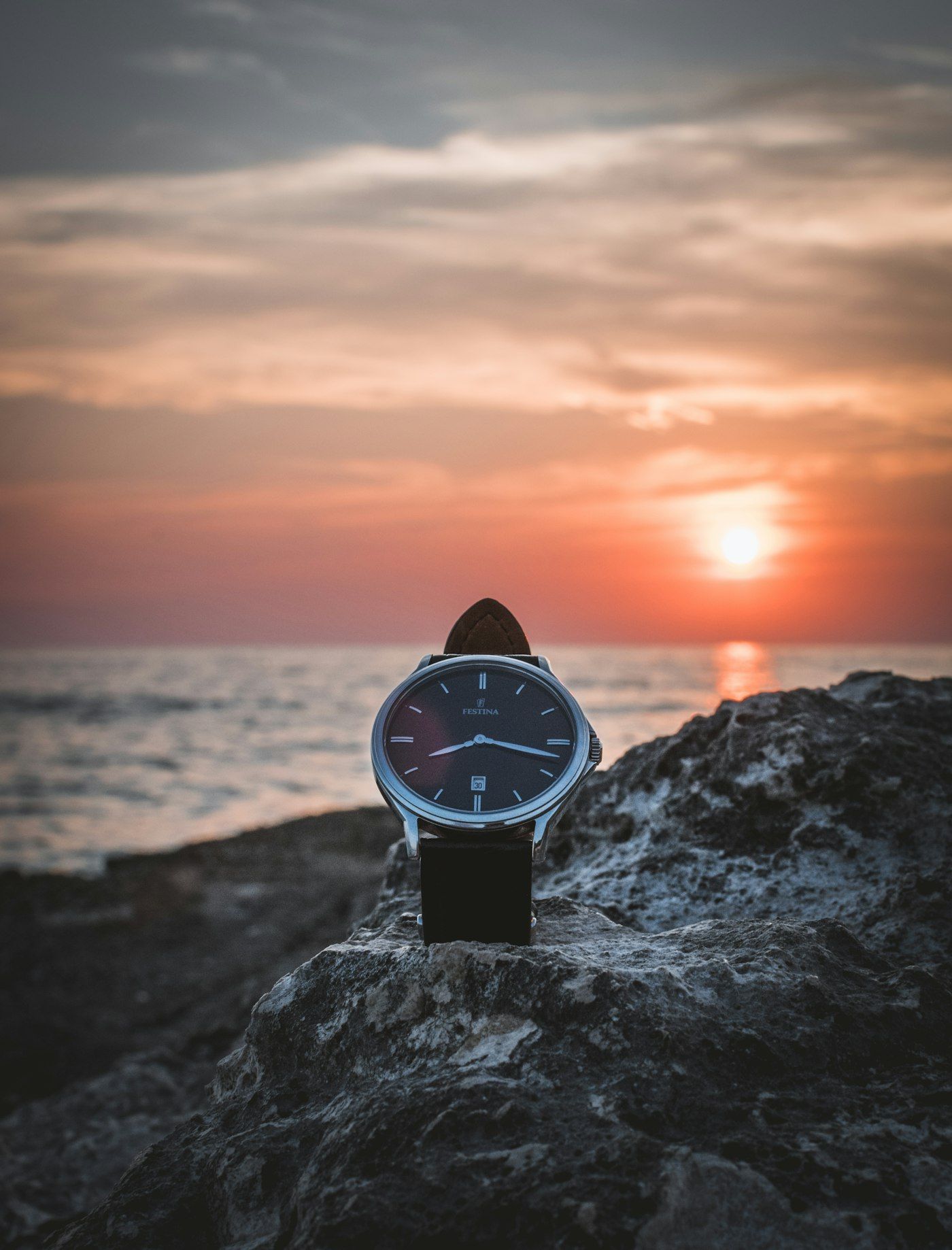 A wristwatch resting on a rock with the sea and a low sun in the background.