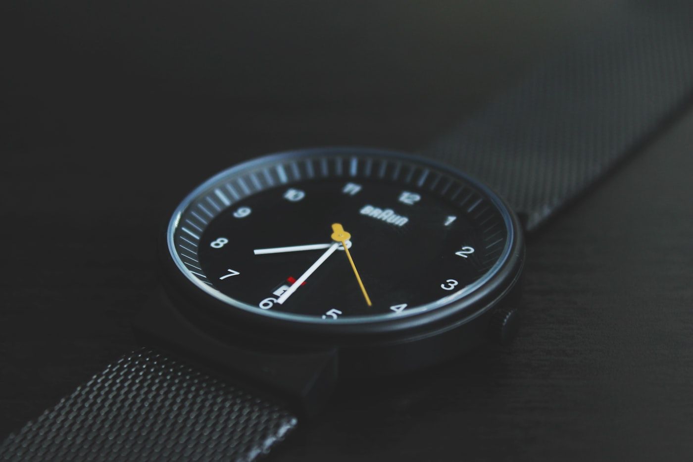 A wristwatch with a dark dial fitted to a woven steel mesh strap, close up.